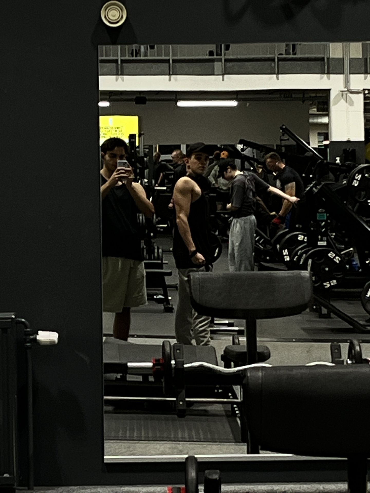 Gym mirror reflection of a person taking a photo, with equipment and people exercising in the background.